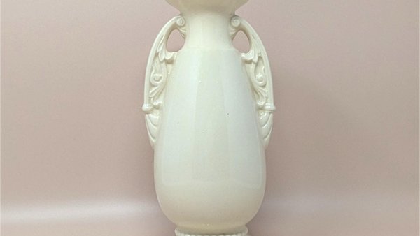 A studio photo of a tall, cream-colored ceramic vase with a glossy finish. The vase features an elegant urn shape with two ornate, scroll-style handles on the sides. It has a flared rim and sits on a tiered circular base with a beaded detail around the bottom.