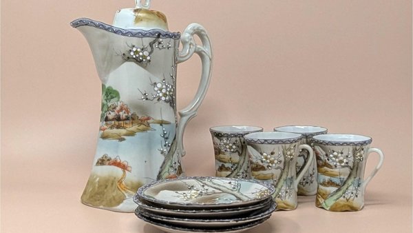 A hand-painted porcelain tea set featuring a tall teapot, four cups, and four saucers. The set depicts a traditional Japanese landscape with mountains, water, and flowering trees in soft earth tones and blue borders.